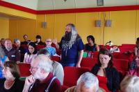 A man with long hair and beard stands in the midst of an audience, asking a question